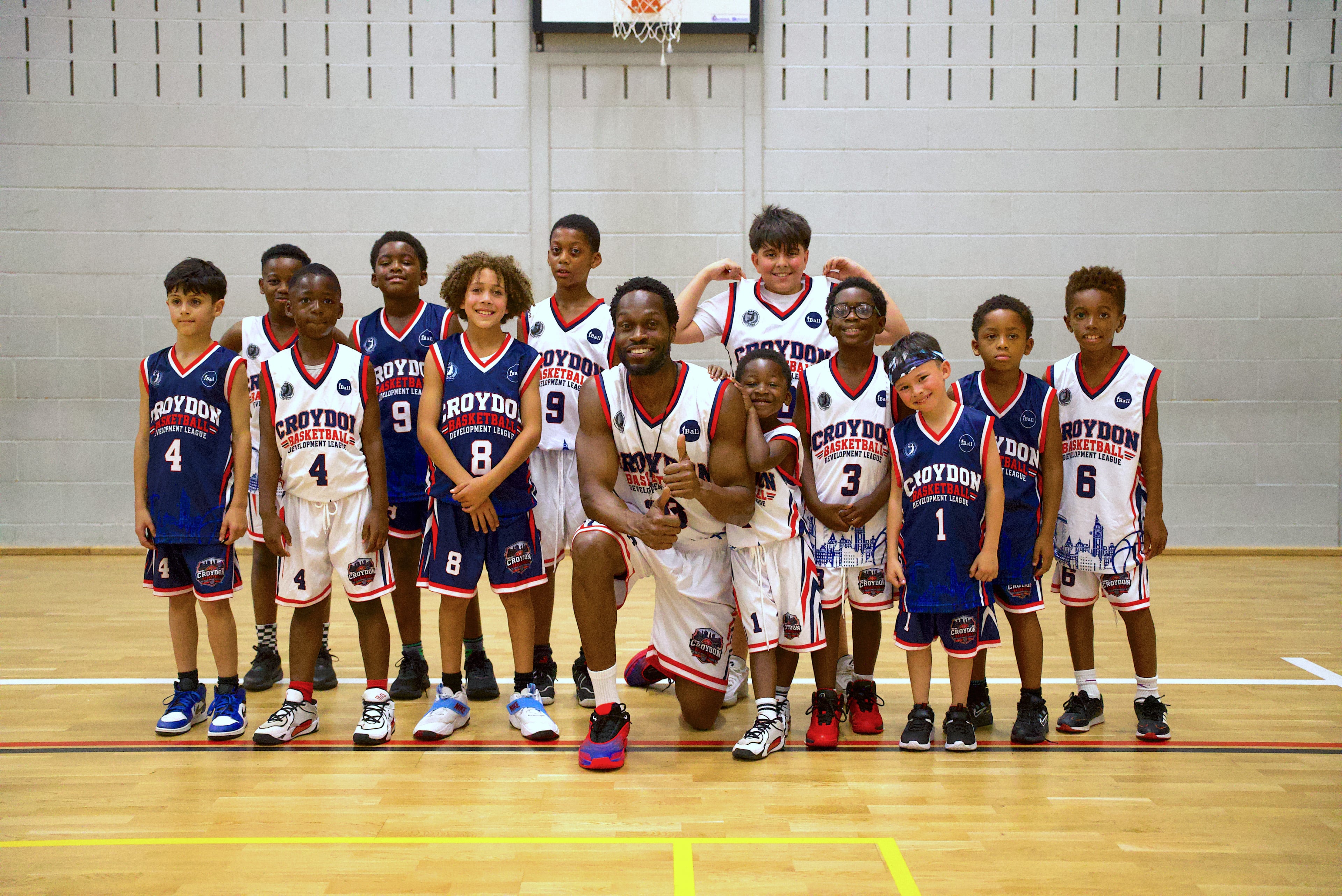 A smiling iBall coach kneeling with a group of young players in Croydon Basketball Development League kit